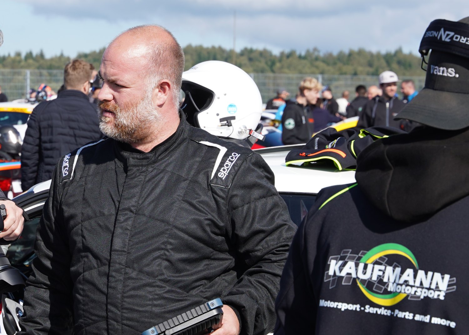 Christian Kaufmann im Rennanzug im Fahrerlager, im Hintergrund Fahrzeuge, Helme und weitere Teilnehmer