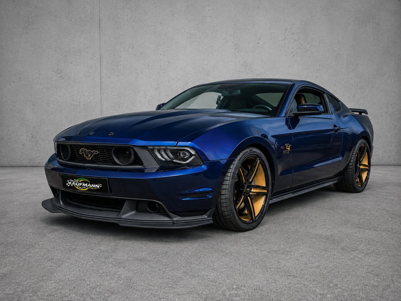 Blauer Ford Mustang mit goldenen Felgen und Kaufmann-Motorsport-Kennzeichenhalter vor einer Betonwand