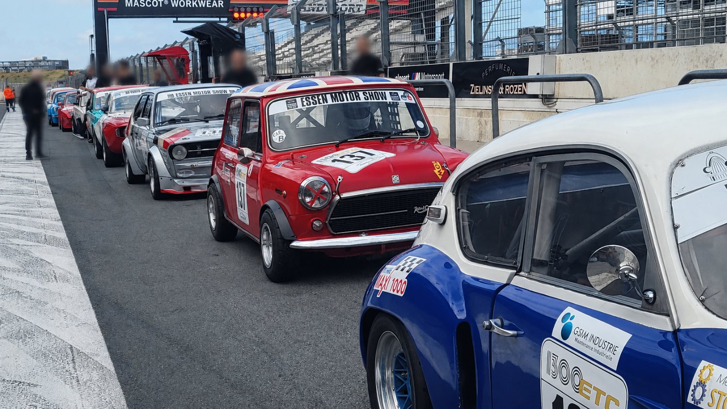 Roter Mini mit Startnummer 131 in der Boxengasse, davor und dahinter weitere klassische Rennfahrzeuge