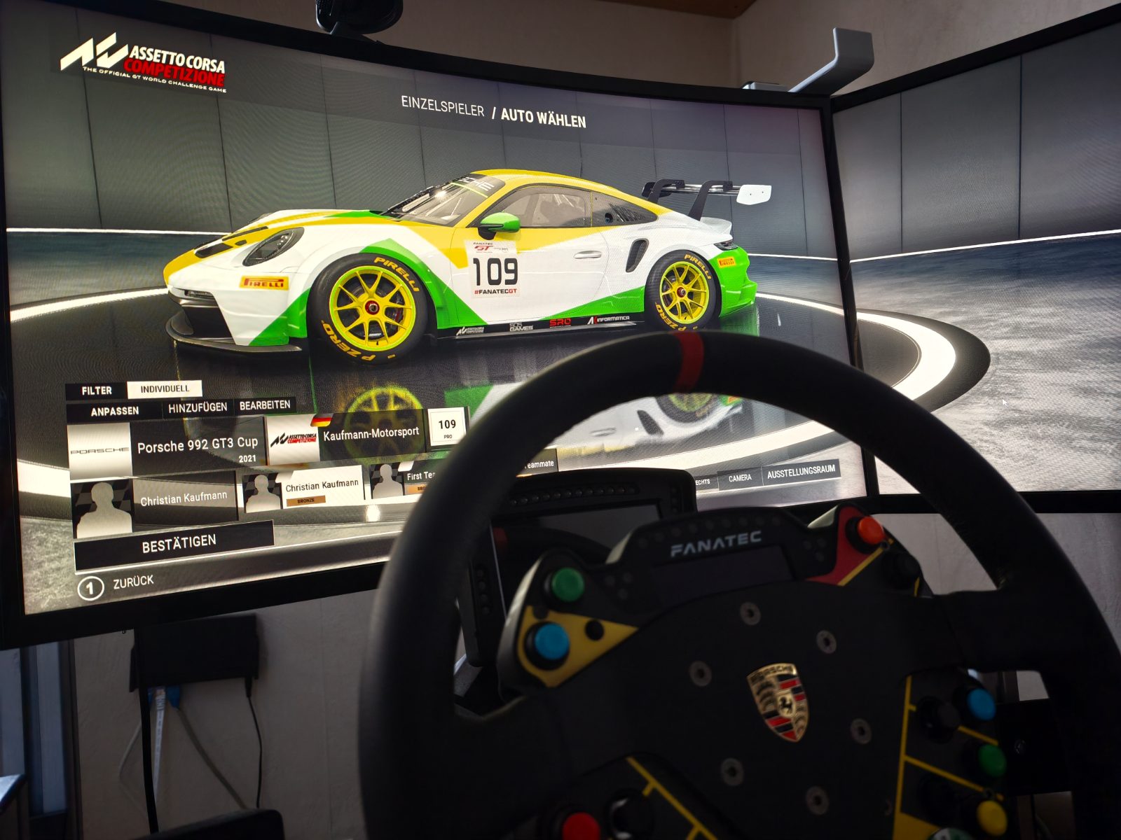 Das SimRacing-Rig mit Lenkrad vor dem gebogenen Bildschirm, im Spiel ein Fahrzeug in den Farben von Kaufmann Motorsport