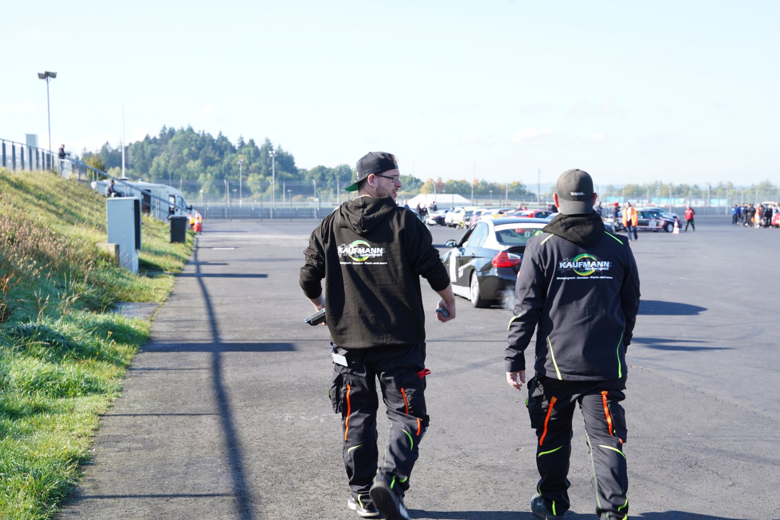 Zwei Mitarbeiter in Kaufmann-Motorsport-Hoodies gehen durch das Fahrerlager in Richtung der Fahrzeuge